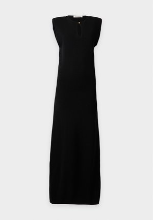 Personne grande portant une robe maxi noire sans manches avec un décolleté en trou de serrure à l'avant et des chaussures noires pointues, debout devant un fond neutre.