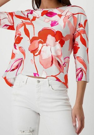 Vrouw draagt witte broek en een witte top met lange mouwen, met grote rode en roze bloemenpatronen, kort boven de taille.