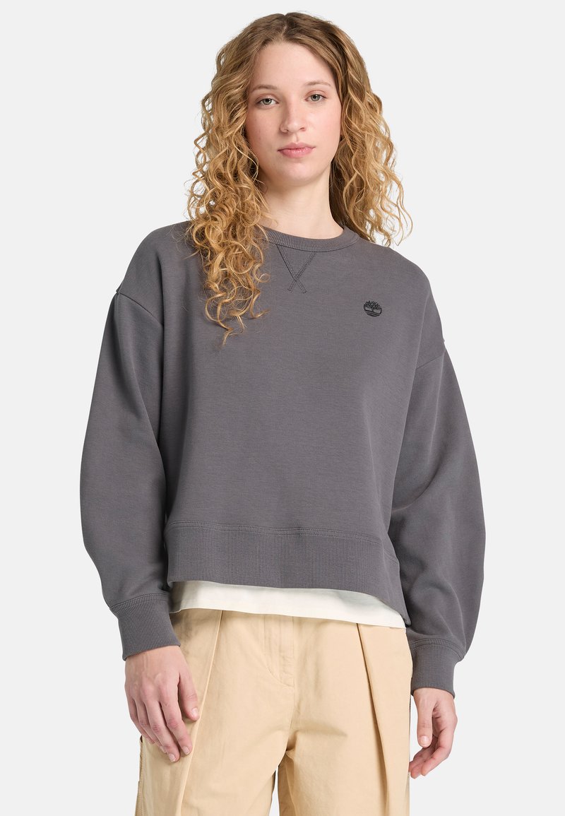 Sweat-shirt gris avec manches surdimensionnées, encolure ronde, poignets et ourlet côtelés. Présente un petit logo noir sur la poitrine. Porté par-dessus un haut blanc.