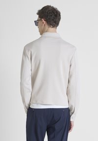 Pulóver de color beige claro con cuello, presenta una textura suave y sutiles detalles de costura. Se lleva sobre una camisa blanca y unos pantalones azul marino.