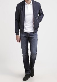 Marinblå bomberjacka över en vit polo, parat med mörk denimjeans och svarta kängor. Modellen står med händerna i fickorna.