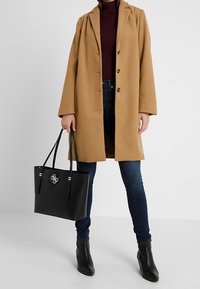 Karmelowy płaszcz z wełny z dużymi klapami i zapięciem na guziki, w połączeniu z ciemnymi dżinsami i czarną torbą na ramię z metalicznym logo i akcentami.