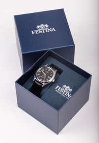 Melns analogs pulkstenis ar baltām ciparnīcām, sudraba korpuss un melna ādas siksniņa, pasniegts tumši zilā kastē ar mīkstu iekšpusi.