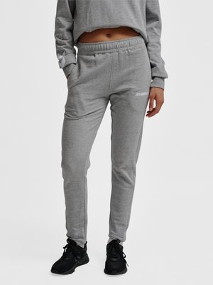 Personne portant un pantalon de survêtement gris avec une ceinture élastique et des poches latérales, associée à des chaussures de sport noires, debout devant un fond uni.