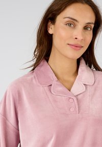 Top de pana rosa con cuello y dos cierres de botón. Presenta una textura suave y detalles de costura en el escote.