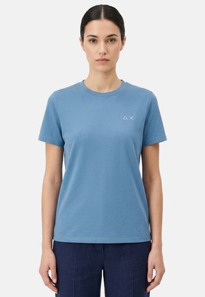 Donna con capelli scuri raccolti, indossa una maglietta blu semplice a maniche corte e pantaloni scuri, in piedi davanti a uno sfondo chiaro uniforme.