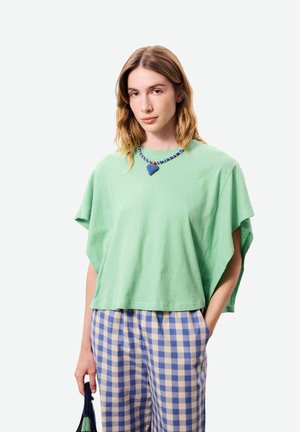 Jeune femme portant un haut ample vert menthe, un pantalon à carreaux bleu et blanc, tenant un sac à main bleu marine et portant un collier avec un pendentif en forme de cœur bleu.