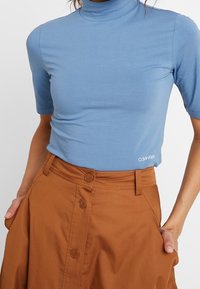 Vaaleansininen, lyhythihainen poolopaita, joka on valmistettu pehmeästä kankaasta ja jossa on hienovarainen Calvin Klein -logo helmassa, yhdistettynä ruskeaan nappikiinnityksellä varustettuun hameeseen.