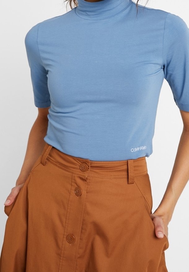 Vaaleansininen, lyhythihainen poolopaita, joka on valmistettu pehmeästä kankaasta ja jossa on hienovarainen Calvin Klein -logo helmassa, yhdistettynä ruskeaan nappikiinnityksellä varustettuun hameeseen.