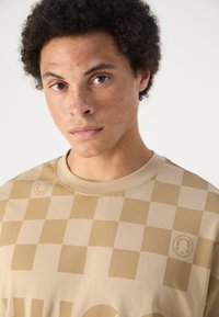 Camiseta de algodón color beige con un patrón a cuadros, con cuello redondo y un pequeño detalle de logo. Textura suave y corte relajado.