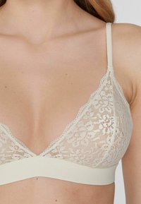 Krajkový bralet v světlé béžové barvě s květinovým vzorem, s trojúhelníkovými košíčky, tenkými ramínky a pevným páskem pod prsy.