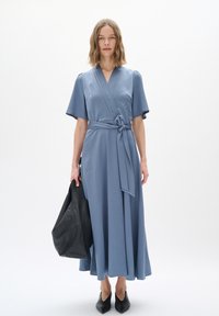 Blaues Wickelkleid aus strukturiertem Stoff, mit kurzen Ärmeln, einem Taillengürtel und einem schwingenden Rock. Kombiniert mit einer großen schwarzen Tragetasche.