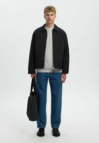 Veste noire zippée sur un sweat gris, assortie à un jean bleu. Le mannequin tient un grand sac noir. L'arrière-plan est blanc uni.