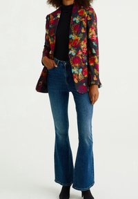 Květinové sako z tmavé látky s výraznými červenými, růžovými a tyrkysovými květy, doplněné středně vysokými modrými rozšířenými džínami a černými botami.