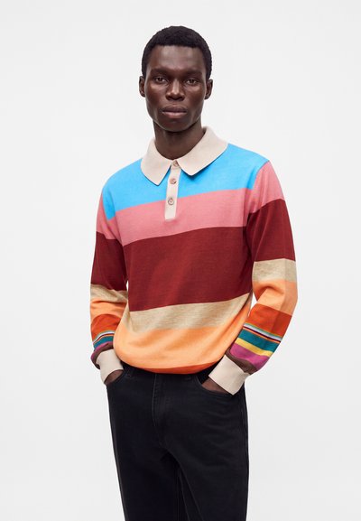 Homme portant un polo à manches longues rayé multicolore, les mains dans les poches d'un pantalon noir, sur un fond uni.