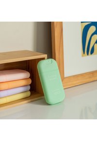 Bottiglia di detergente per le mani verde etichettata "Verbena Purificante", posizionata accanto a bottiglie color pastello su uno scaffale di legno. Superficie liscia, bordi arrotondati.