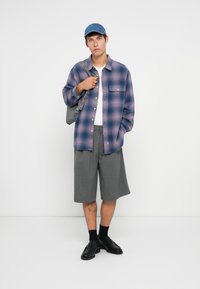Checked flannel shirt in pink and blue, gray shorts, black loafers, and a denim cap, holding a gray shoulder bag. Smooth textures and casual design.