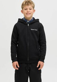 Črna zip-up kapuca z sprednjim žepom, pletenimi manšetnimi rokavi in kapuco; na levi strani prsnega predela ima bele vezenine. Oblečena z karirastim srajcem.