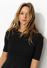 Robe noire ajustée à manches courtes, présentant une texture lisse et une coupe subtile, portée par une personne ayant de longs cheveux ondulés devant un fond uni.