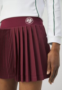 Burgundy plisserad kjol med ett brett midjeband som visar en "Roland Garros"-logotyp, tillverkad av texturerat tyg, som framhäver skarpa veck och en ren finish.