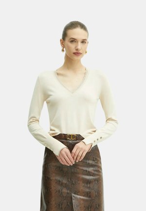 Maglione beige con scollo a V e maniche lunghe, caratterizzato da bordi texturizzati e dettagli con bottoni sui polsini. Abbinato a una gonna aderente con motivo pitone marrone.