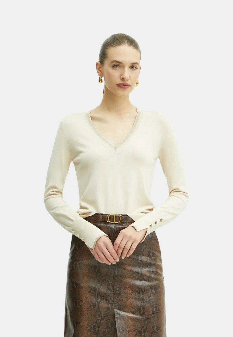 Maglione beige con scollo a V e maniche lunghe, caratterizzato da bordi texturizzati e dettagli con bottoni sui polsini. Abbinato a una gonna aderente con motivo pitone marrone.