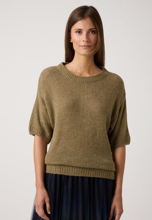 Eine Frau trägt einen lockeren olivgrünen Strickpullover mit Ellenbogenlangen Ärmeln und einen marineblauen plissierten Rock. Sie steht vor einem einfachen hellen Hintergrund.