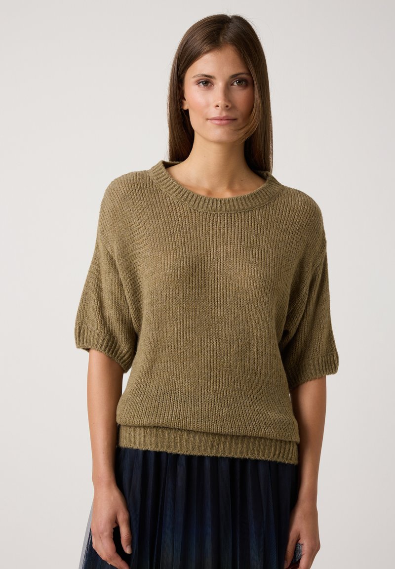 Eine Frau trägt einen lockeren olivgrünen Strickpullover mit Ellenbogenlangen Ärmeln und einen marineblauen plissierten Rock. Sie steht vor einem einfachen hellen Hintergrund.