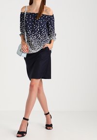 Marin off-shoulder topp med vita prickar, kombinerad med en svart kjol. Svarta klacksandaler och en ljusblå crossbody-väska fulländar looken.