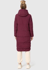 Lång maroon puffjacka med huva, med horisontell sömnad, avslappnad passform och längd under knäna. Bärs över ljusa byxor.