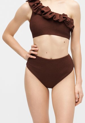 Femeie purtând un costum de baie din două piese, de culoare maro închis texturat, cu top asimetric cu volane pe un umăr și pantaloni scurți cu talie înaltă, pe un fundal neutru.