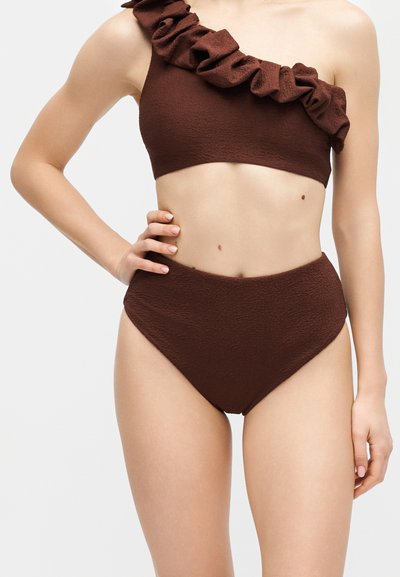 Femme portant un maillot de bain deux-pièces brun foncé texturé avec un haut asymétrique à volants et un bas taille haute, sur un fond neutre.