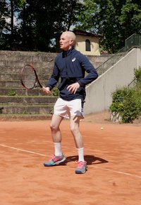 Tennista in giacca blu scuro e pantaloncini bianchi, che tiene una racchetta rossa, indossando sneakers blu con accenti rosa su un campo in terra rossa.