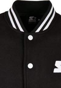 Zwarte varsityjack met een gestreepte witte kraag, voorzien van witte drukknopen en een geborduurd logo op de voorkant. Zachte stoftextuur.
