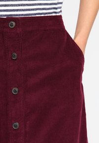 Burgundy kordflortkjol med knäppning framtill, med vertikala ribbor, sidofickor och en mjuk textur, kombinerad med en randig skjorta.