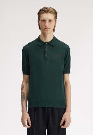 Junger Mann mit kurzem blondem Haar trägt ein dunkelgrünes kurzärmliges Polo mit rotem Lorbeer-Logo und schwarzer Hose, steht vor einfarbigem Hintergrund.