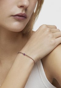 Pulsera de cuentas doradas y moradas con una gema morada central, llevada en una muñeca con una textura de piel suave y clara.