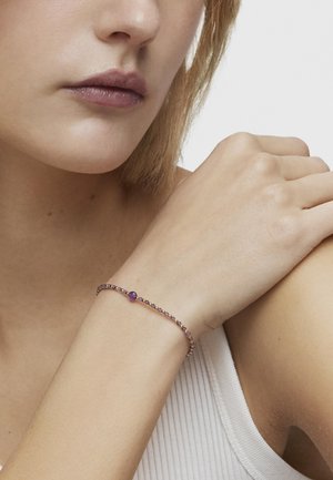 Bracciale di perline dorate e viola con una pietra preziosa centrale viola, indossato su un polso con una texture della pelle liscia e chiara.