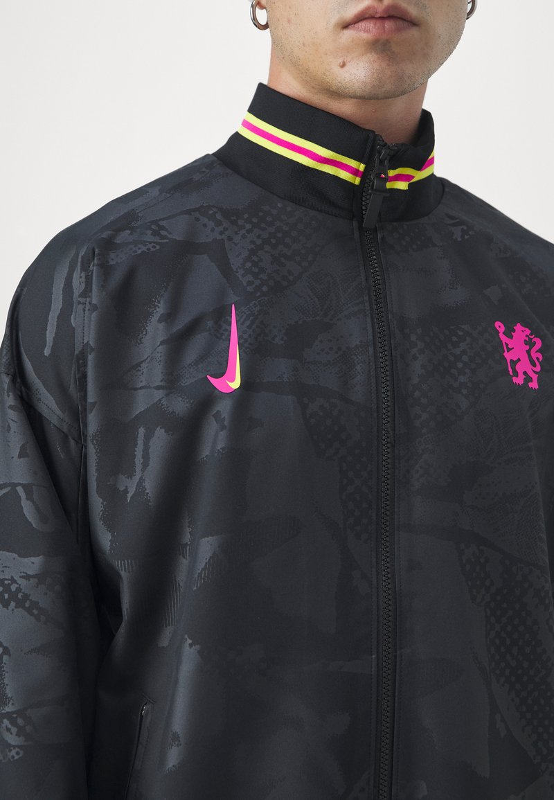 Giacca nera con zip e motivo testurizzato, caratterizzata da un Swoosh Nike rosa e uno stemma. Il colletto presenta dettagli a strisce gialle e rosa.