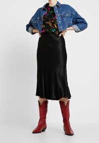 Denimjacke, schwarzes Top mit Blumenmuster, schwarzer Satinrock und rote Cowboystiefel mit spitzen Zehen. Fokus auf Texturen und Farbkontraste.