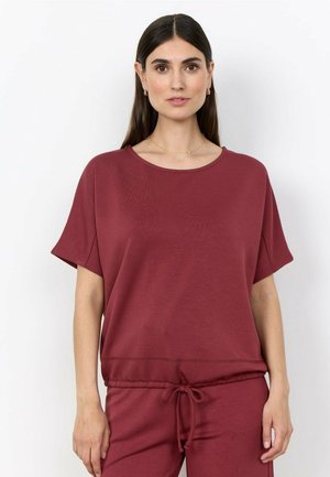 Vrouw met steil donker haar draagt een bordeauxkleurige top met korte mouwen en bijpassende broek, staande tegen een effen witte achtergrond.