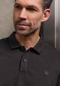 Homme à la barbe courte portant un polo noir à col avec des boutons et un petit logo circulaire sur la poitrine.