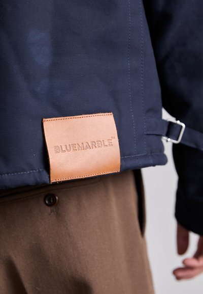 Navy waterproof jacket with a brown leather label attached, featuring textured fabric, a sleek design, and a silver buckle detail.