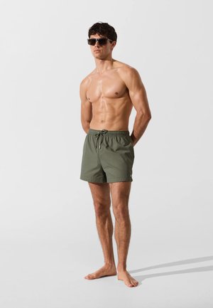 Hombre sin camiseta con pantalones cortos de baño verde oscuro y gafas de sol negras, de pie descalzo sobre un fondo claro y llano.
