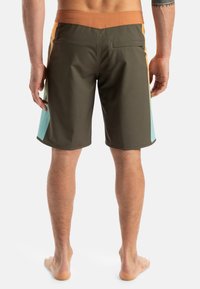 Simshorts med en olivgrön kropp och sidan paneler i orange och aquablå, en brun midja och en bakficka. Slät textur.