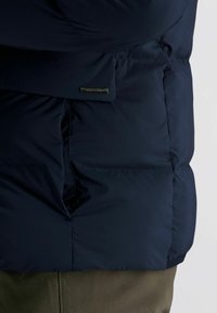 Close-up van een marineblauwe gewatteerde jas met een rechthoekig metalen merketiket op de zakflap, gedragen over olijfgroene broek.