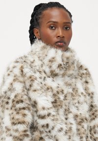 Brun og hvit faux fur-kappe med høy krage, med et prikket mønster og en myk, teksturert overflate.