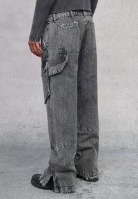 Pantalones cargo de pierna ancha en denim desgastado gris, con múltiples bolsillos, dobladillos deshilachados y un ajuste relajado. Los acentos incluyen una superficie texturizada.