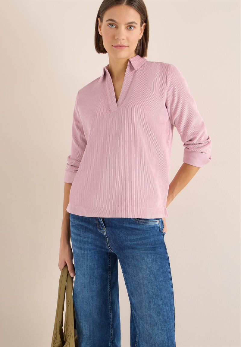 Top texturizado en rosa claro, con cuello y escote en V, y mangas enrolladas, combinado con jeans de pierna ancha azules. Diseño informal adecuado para el uso diario.