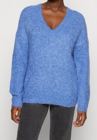 Blå, fluffig V-ringad tröja som bärs tillsammans med mörkgrå byxor, visar överkropp och armar av en person med mörk hudton.